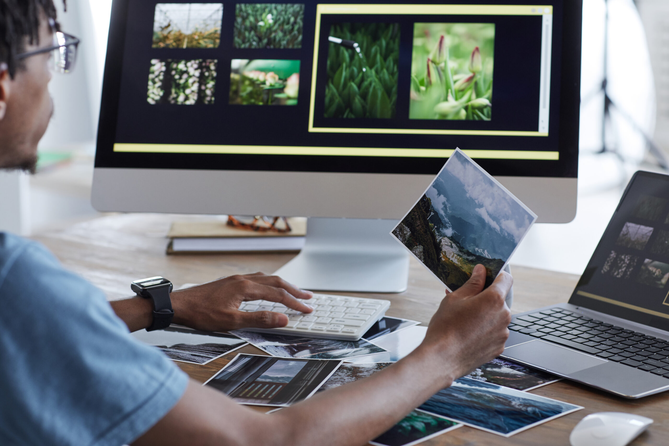 Back view portrait of young African-American man holding printed photographs while using editing software via computer while working at desk in home office, copy space