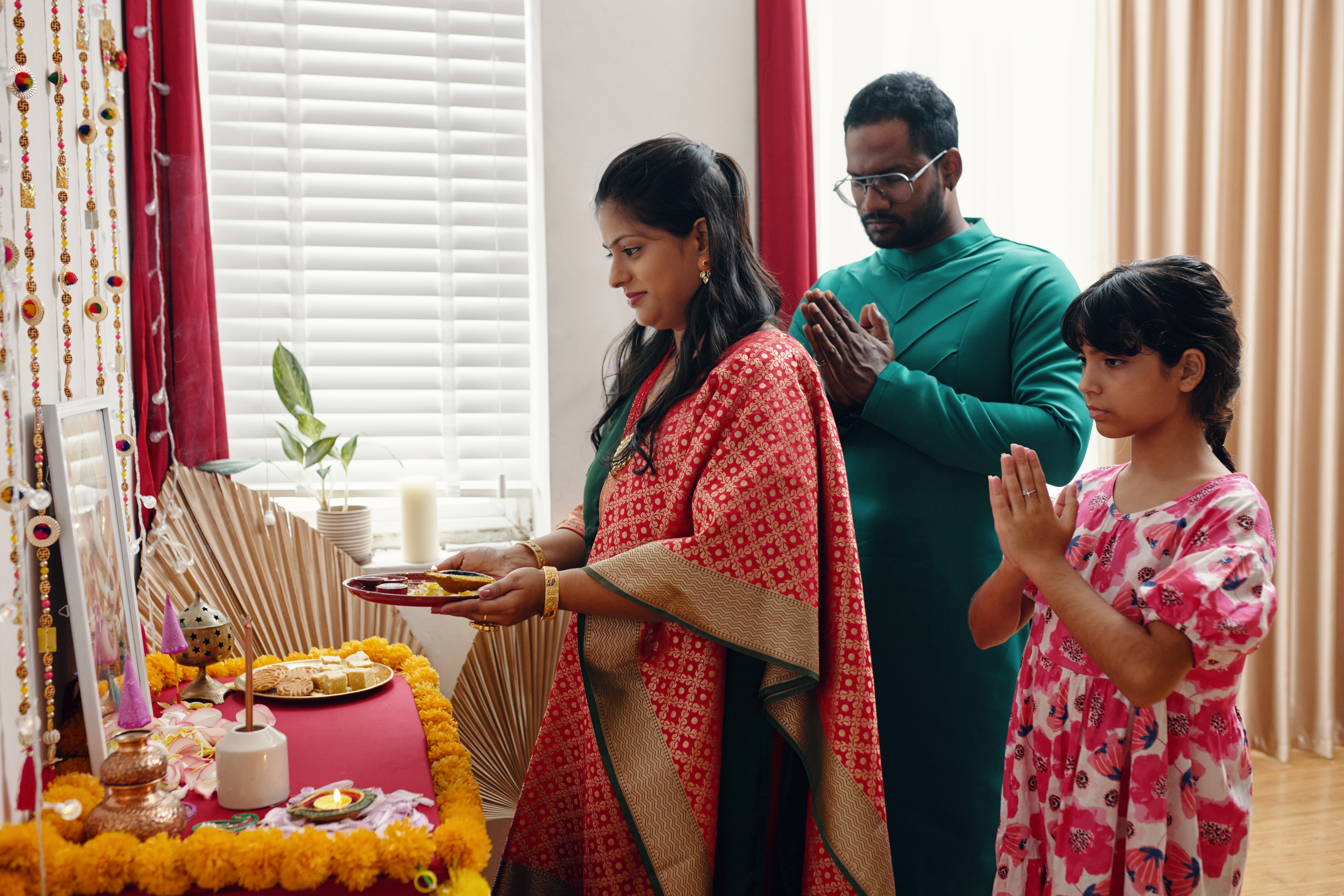 Family participating in traditional cultural ritual, standing in prayer. Family members are dressed in colorful attire, with offerings and decorative items on altar table