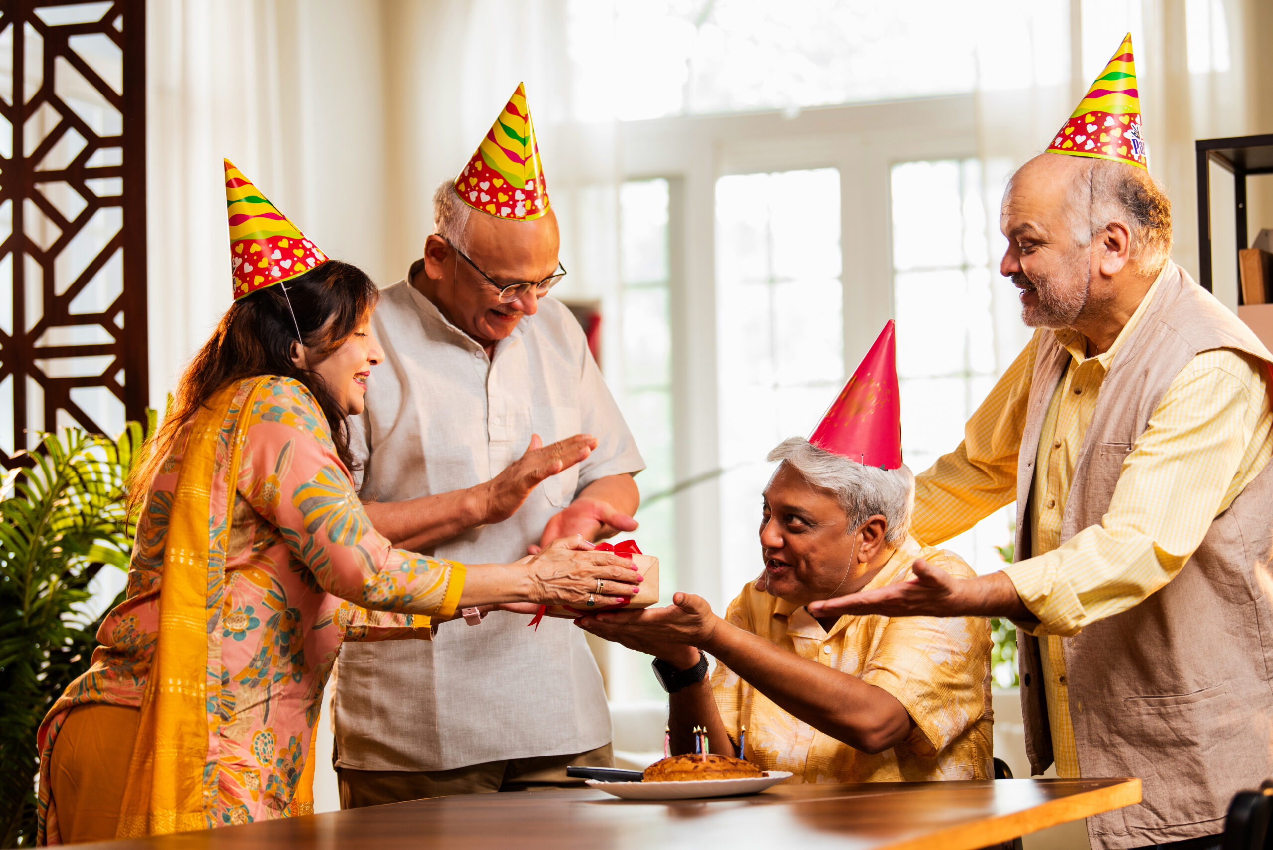 Indian senior friends celebrating birthday at home, cutting cake, giving receiving gifts with laughter, wearing party hats, surrounded by candles, cheerful Asian elders enjoying reunion celebration